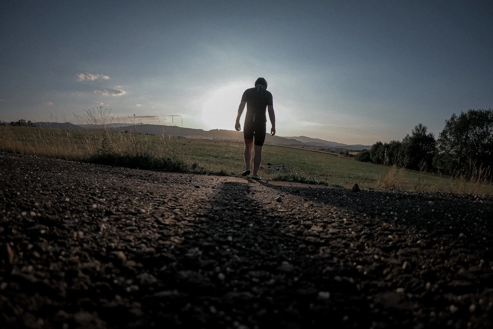 Sylwetka kolarza od tyłu idącego w stronę zachodu słońca, rower leży na trawie przed nim